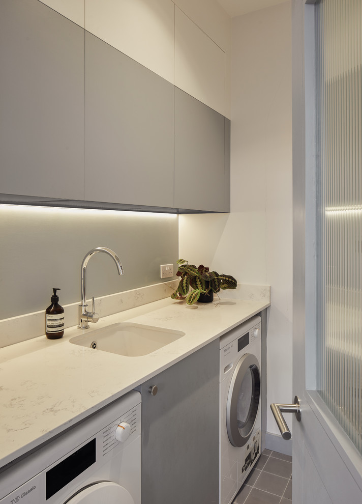 Scandinavian single-wall dedicated laundry room in London with an undermount sink, flat-panel cabinets, grey cabinets, white walls, a side-by-side washer and dryer, grey floor and white benchtop.