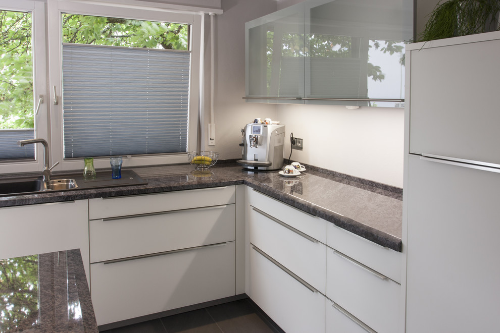 This is an example of a mid-sized contemporary galley open plan kitchen in Stuttgart with a drop-in sink, flat-panel cabinets, white cabinets, granite benchtops, white splashback, stone tile splashback, black appliances, ceramic floors, a peninsula, grey floor and grey benchtop.