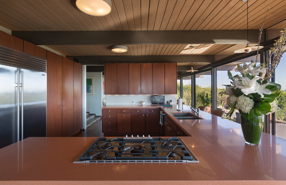 Mid-sized midcentury l-shaped open plan kitchen in Los Angeles with an undermount sink, flat-panel cabinets, dark wood cabinets, solid surface benchtops, stainless steel appliances, dark hardwood floors, a peninsula and brown benchtop.