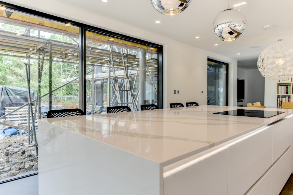 Photo of a large modern grey and white single-wall kitchen/diner in Sussex with a double-bowl sink, flat-panel cabinets, white cabinets, quartz worktops, multi-coloured splashback, mirror splashback, black appliances, porcelain flooring, an island, grey floors, white worktops and feature lighting.