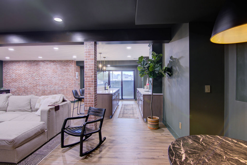 This is an example of a large modern walk-out basement in Kansas City with a home bar, green walls, vinyl flooring, exposed beams and brick walls.
