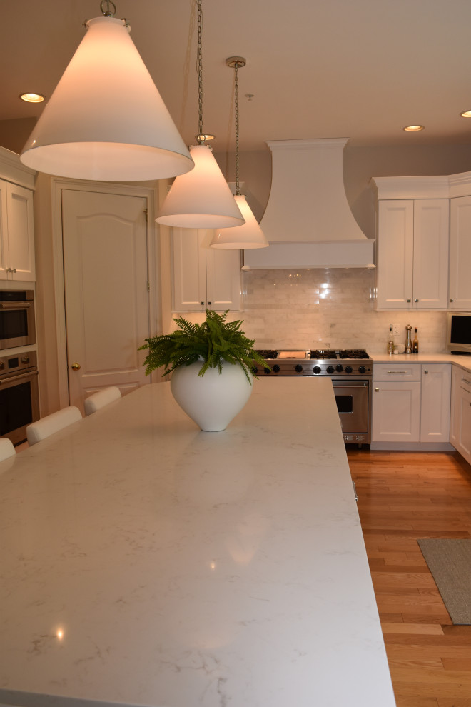 Klassisk inredning av ett stort vit vitt kök, med en undermonterad diskho, luckor med infälld panel, vita skåp, bänkskiva i kvartsit, vitt stänkskydd, stänkskydd i tunnelbanekakel, rostfria vitvaror, ljust trägolv, en köksö och brunt golv