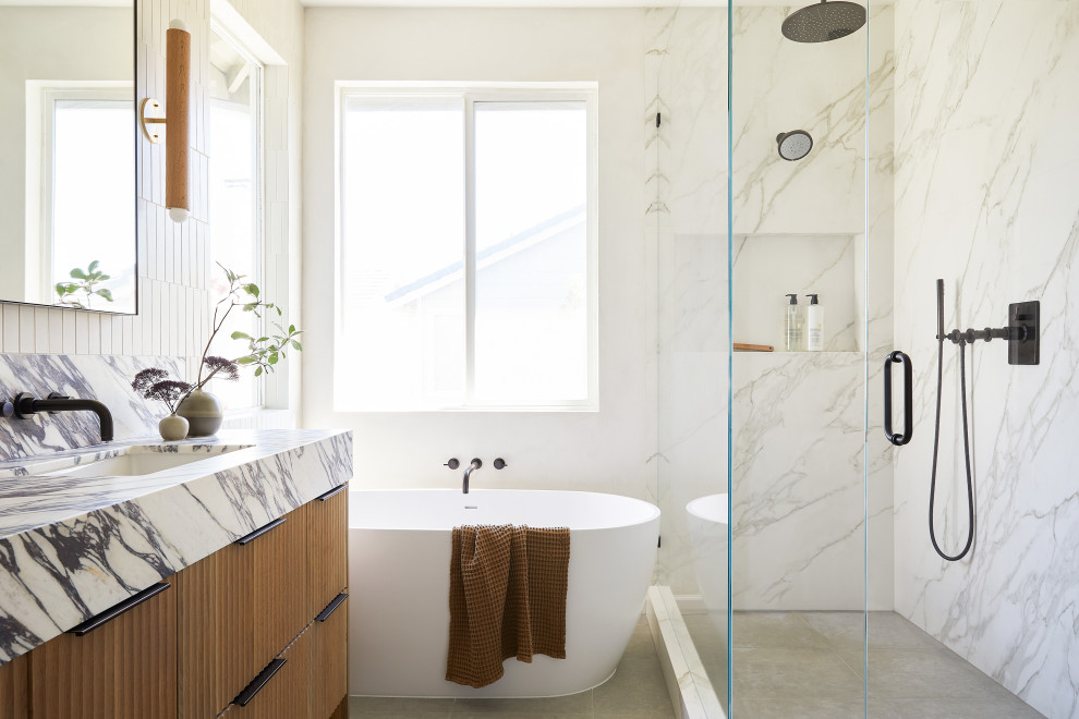 Modern inredning av ett stort lila lila en-suite badrum, med möbel-liknande, skåp i ljust trä, ett fristående badkar, en hörndusch, en toalettstol med separat cisternkåpa, beige kakel, keramikplattor, vita väggar, kalkstensgolv, ett undermonterad handfat, marmorbänkskiva, grått golv och dusch med gångjärnsdörr