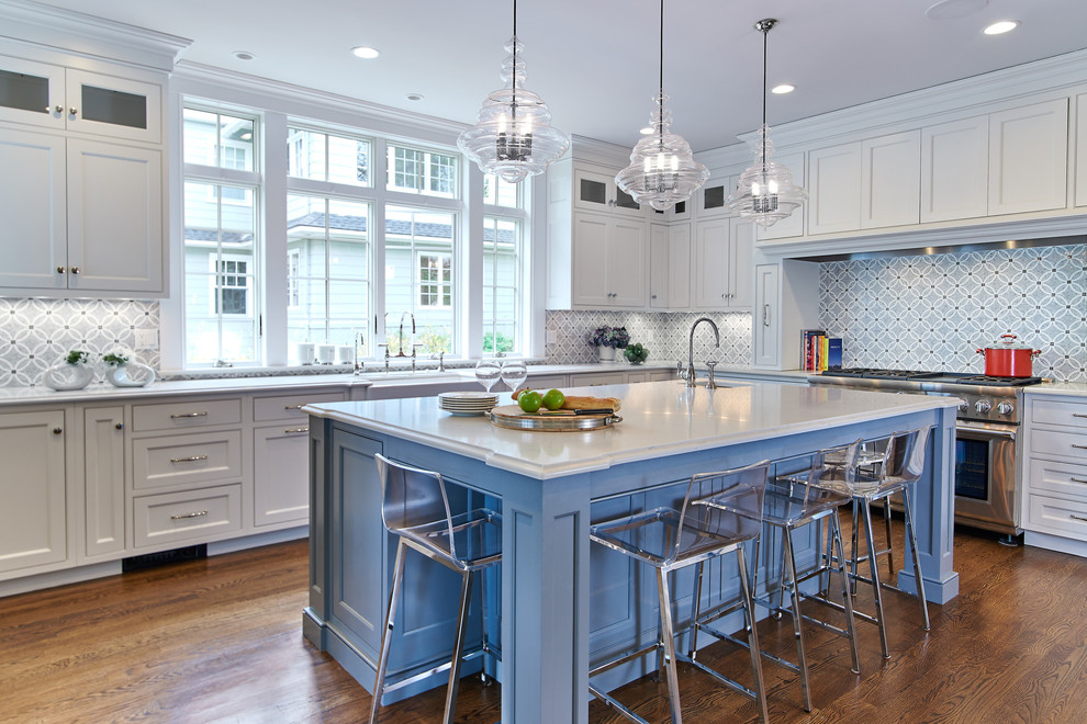Beach style l-shaped kitchen in New York with a farmhouse sink, shaker cabinets, white cabinets, multi-coloured splashback, stainless steel appliances, medium hardwood floors, with island and white benchtop.