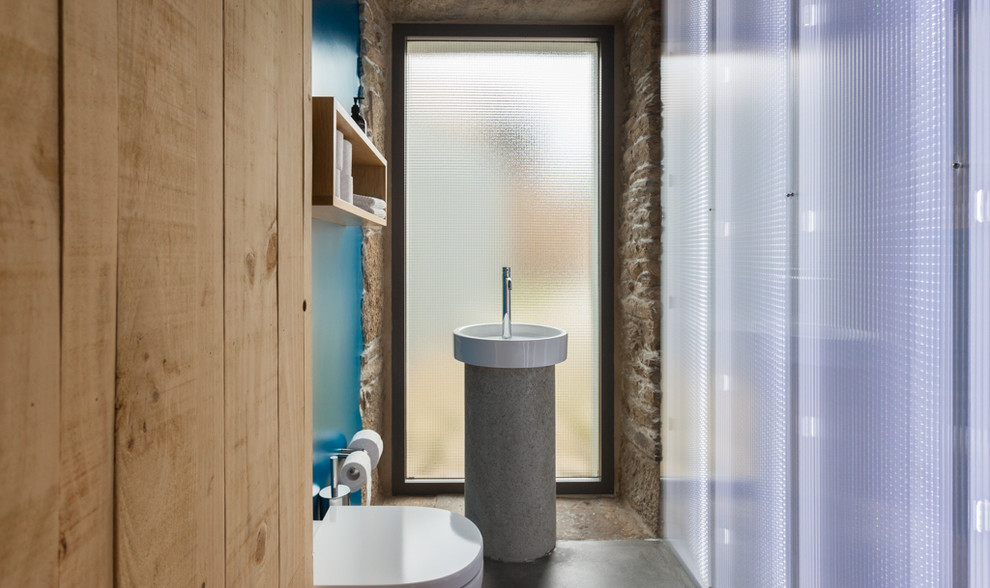 Photo of a small contemporary 3/4 bathroom in Other with open cabinets, light wood cabinets, a wall-mount toilet, blue walls, concrete floors and a pedestal sink.