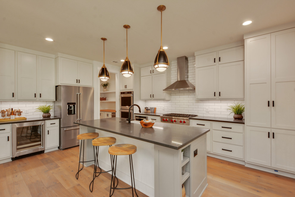 This is an example of a transitional l-shaped eat-in kitchen in Austin with a farmhouse sink, recessed-panel cabinets, grey cabinets, white splashback, subway tile splashback, stainless steel appliances, light hardwood floors, with island, brown floor and grey benchtop.