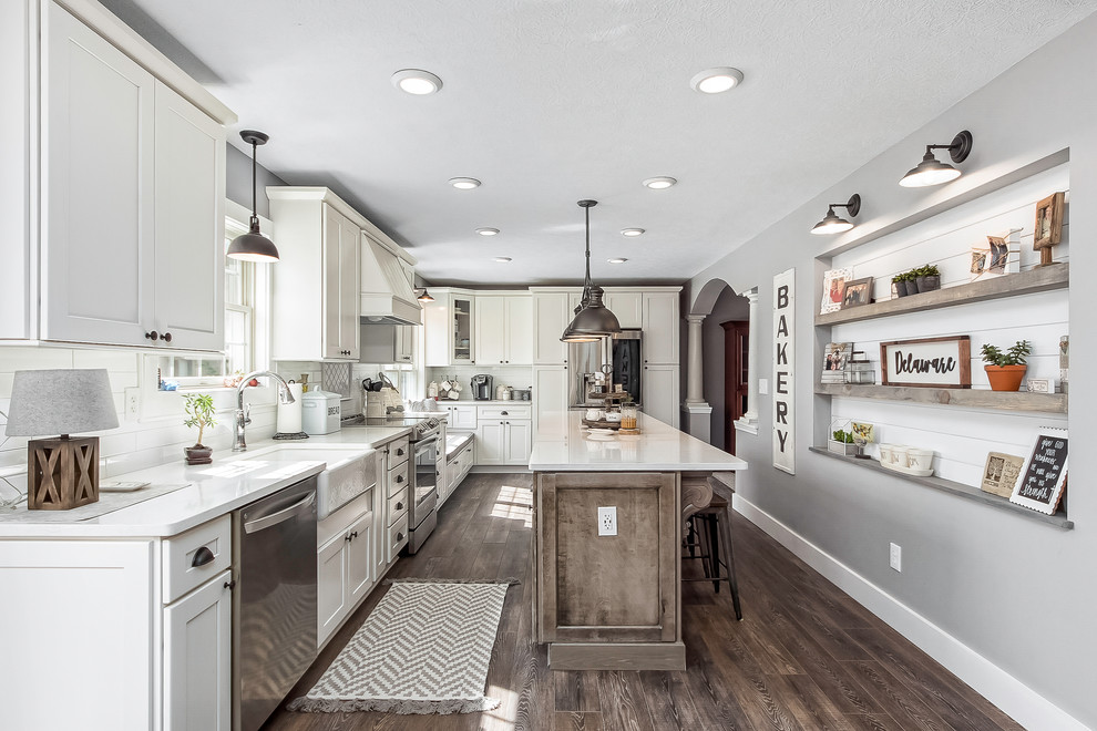 Large country l-shaped kitchen in Columbus with a farmhouse sink, shaker cabinets, white cabinets, white splashback, subway tile splashback, stainless steel appliances, dark hardwood floors, with island, brown floor, white benchtop and quartz benchtops.