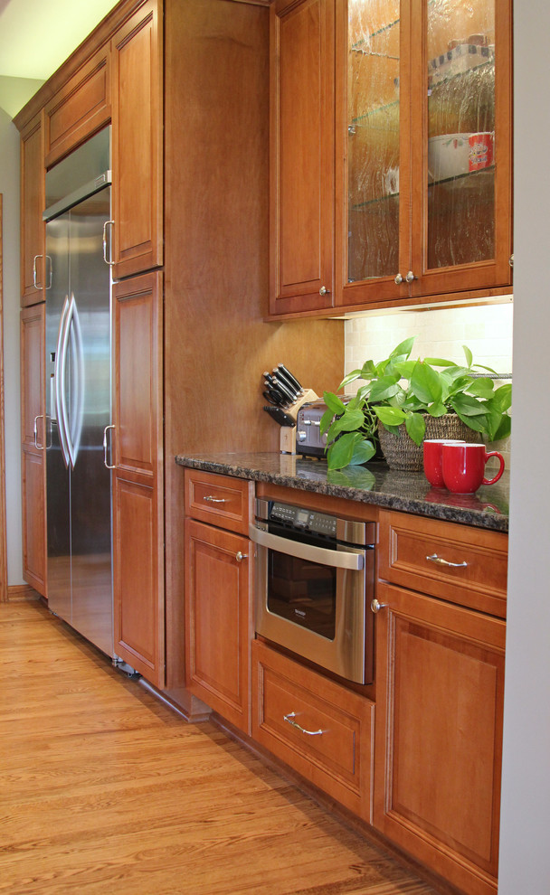 Photo of a large traditional l-shaped open plan kitchen in Milwaukee with an undermount sink, recessed-panel cabinets, brown cabinets, granite benchtops, beige splashback, ceramic splashback, stainless steel appliances, light hardwood floors, multiple islands, brown floor and black benchtop.