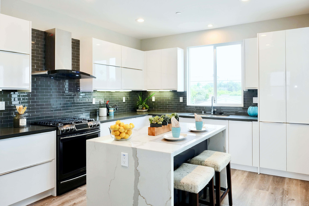 Mid-sized contemporary u-shaped eat-in kitchen in Orange County with a drop-in sink, flat-panel cabinets, white cabinets, quartz benchtops, black splashback, ceramic splashback, black appliances, light hardwood floors, with island, brown floor and black benchtop.