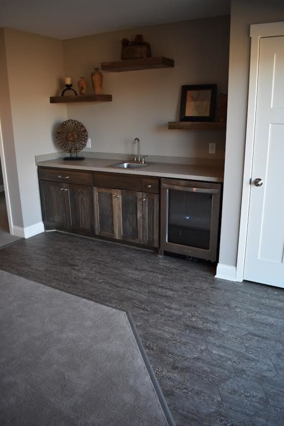 Photo of a small country single-wall wet bar in Minneapolis with a drop-in sink, shaker cabinets, dark wood cabinets, solid surface benchtops, grey splashback, porcelain floors and grey floor.