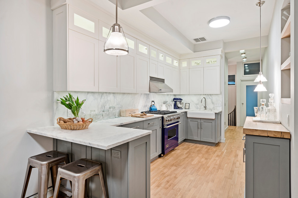This is an example of a mid-sized transitional eat-in kitchen in New York with a farmhouse sink, flat-panel cabinets, grey cabinets, marble benchtops, stone slab splashback, panelled appliances, light hardwood floors and a peninsula.