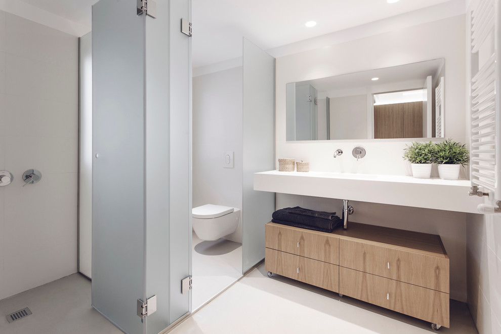 This is an example of a mid-sized powder room in Valencia with a trough sink, flat-panel cabinets, light wood cabinets, white walls and a wall-mount toilet.