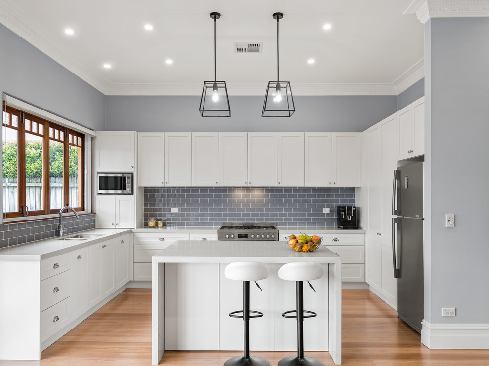 This is an example of a mid-sized transitional u-shaped open plan kitchen in Sydney with an undermount sink, shaker cabinets, white cabinets, quartz benchtops, grey splashback, ceramic splashback, stainless steel appliances, light hardwood floors, with island, brown floor and grey benchtop.