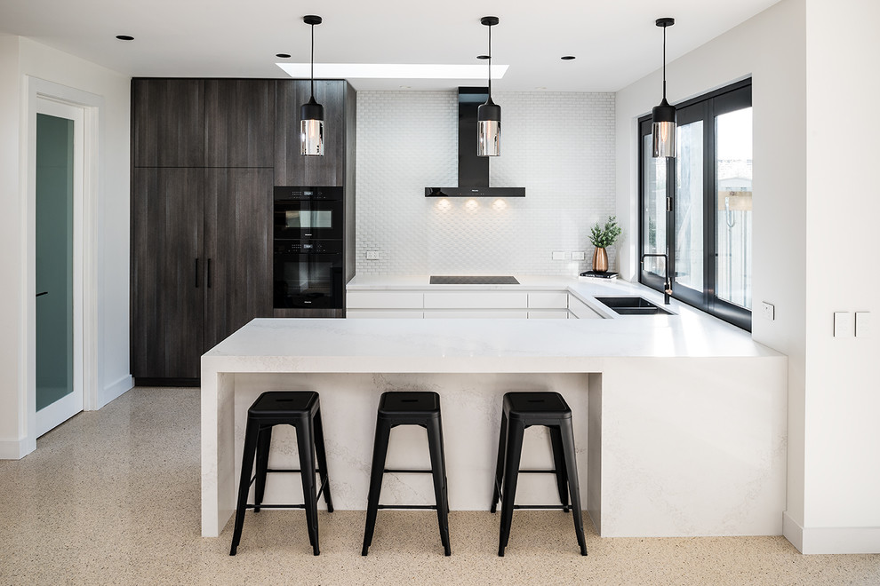 Mid-sized contemporary u-shaped eat-in kitchen in Melbourne with an undermount sink, flat-panel cabinets, white cabinets, quartz benchtops, white splashback, ceramic splashback, black appliances, concrete floors, a peninsula, beige floor and white benchtop.