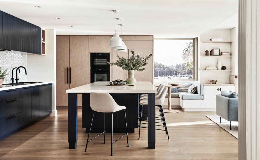 Design ideas for an expansive beach style l-shaped open plan kitchen in Sydney with a drop-in sink, shaker cabinets, blue cabinets, quartz benchtops, green splashback, porcelain splashback, black appliances, light hardwood floors, with island, beige floor and white benchtop.