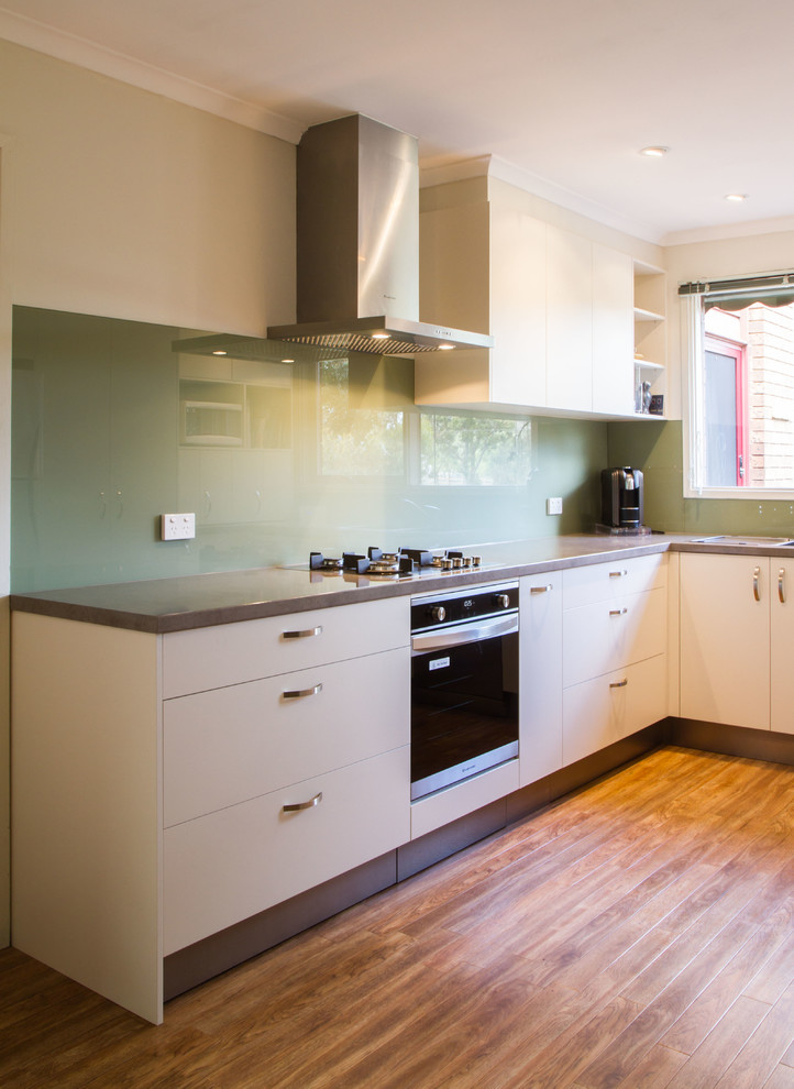 Photo of a small contemporary u-shaped separate kitchen in Melbourne with a drop-in sink, flat-panel cabinets, white cabinets, laminate benchtops, green splashback, glass sheet splashback, medium hardwood floors and no island.
