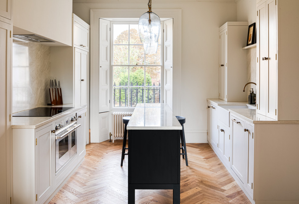 Design ideas for a traditional galley kitchen in Other with a belfast sink, shaker cabinets, white cabinets, stainless steel appliances, medium hardwood flooring, an island, brown floors and beige worktops.