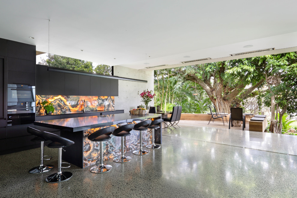 This is an example of a contemporary kitchen in Sunshine Coast with flat-panel cabinets, black cabinets, multi-coloured splashback, black appliances, concrete floors, with island, grey floor and black benchtop.