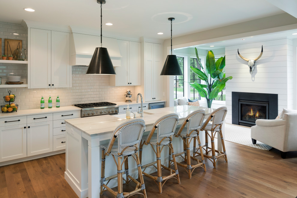 Example of a transitional medium tone wood floor and brown floor kitchen design in Minneapolis with a farmhouse sink, shaker cabinets, white cabinets, white backsplash, subway tile backsplash, stainless steel appliances, an island and white countertops