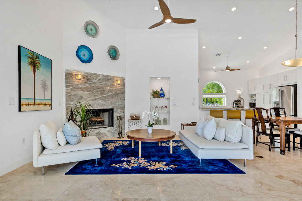 タンパにあるラグジュアリーな中くらいなビーチスタイルのおしゃれなLDK (白い壁、トラバーチンの床、コーナー設置型暖炉、石材の暖炉まわり、ベージュの床、三角天井) の写真