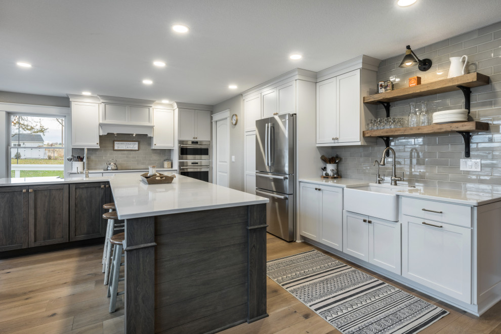 Large country l-shaped medium tone wood floor and brown floor open concept kitchen photo in Boise with a farmhouse sink, shaker cabinets, white cabinets, quartz countertops, gray backsplash, porcelain backsplash, stainless steel appliances, an island and white countertops