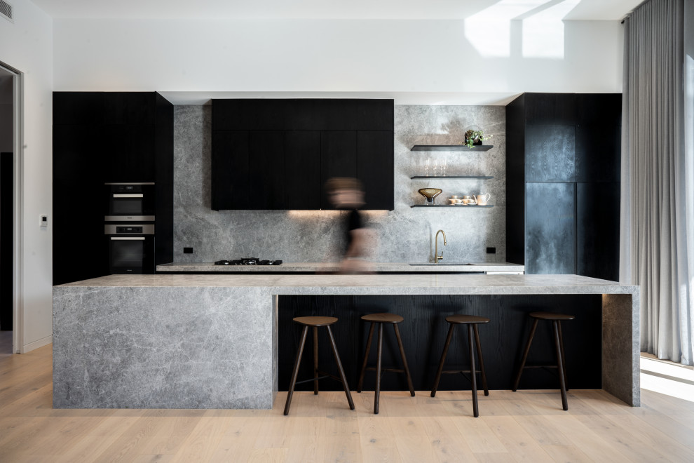 This is an example of a mid-sized contemporary galley kitchen in Melbourne with an undermount sink, flat-panel cabinets, black cabinets, grey splashback, stone slab splashback, black appliances, light hardwood floors, with island, beige floor and grey benchtop.