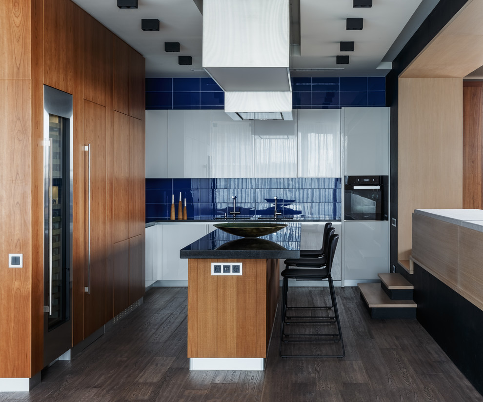 Photo of an expansive contemporary l-shaped open plan kitchen in Moscow with an undermount sink, flat-panel cabinets, white cabinets, granite benchtops, blue splashback, glass tile splashback, black appliances, medium hardwood floors, with island, brown floor and black benchtop.