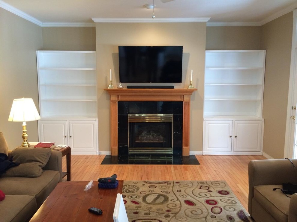 Inspiration for a mid-sized traditional enclosed living room in Burlington with a library, beige walls, light hardwood floors, a standard fireplace, a wood fireplace surround, a wall-mounted tv and beige floor.