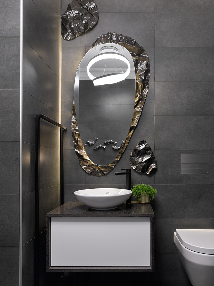 This is an example of a contemporary powder room in Moscow with flat-panel cabinets, white cabinets, a wall-mount toilet, gray tile and a vessel sink.