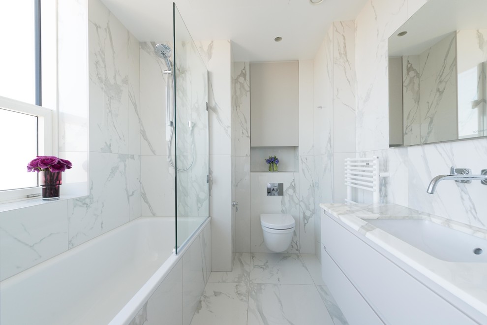 Photo of a mid-sized contemporary bathroom in London with flat-panel cabinets, white cabinets, a drop-in tub, a shower/bathtub combo, a wall-mount toilet, marble, marble floors, a wall-mount sink and white floor.