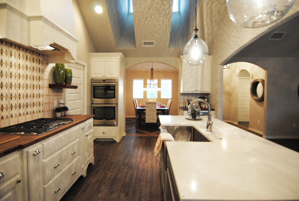 Photo of a mid-sized eclectic u-shaped kitchen in Austin with a farmhouse sink, recessed-panel cabinets, white cabinets, quartz benchtops, beige splashback, ceramic splashback, stainless steel appliances, vinyl floors and with island.