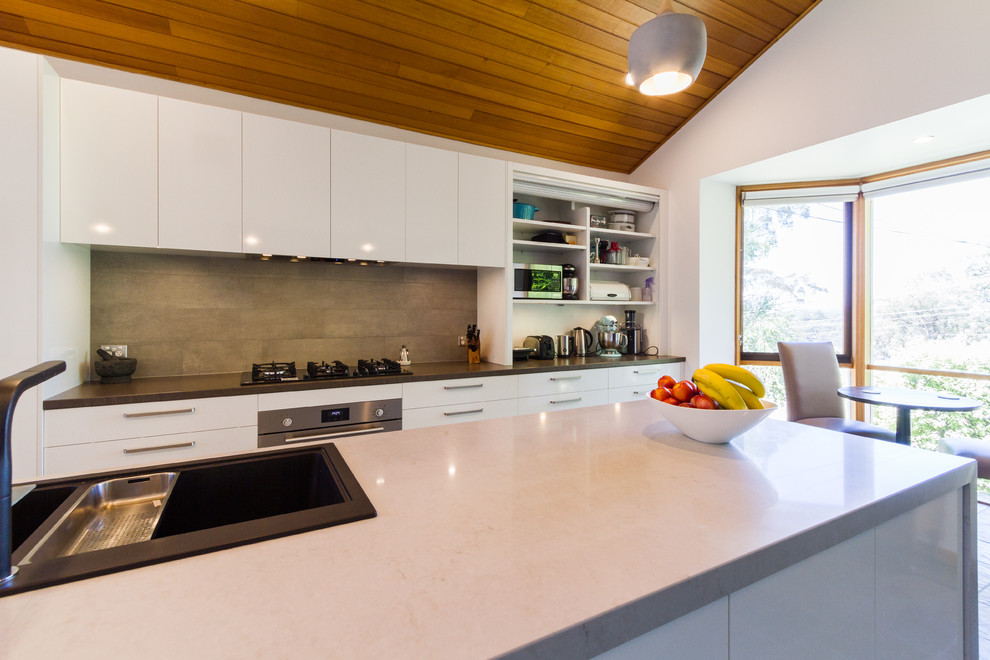 Large midcentury galley eat-in kitchen in Melbourne with a drop-in sink, flat-panel cabinets, white cabinets, quartz benchtops, beige splashback, subway tile splashback, stainless steel appliances, porcelain floors, with island, brown floor and grey benchtop.