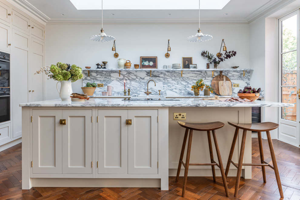 This is an example of a medium sized traditional kitchen/diner in West Midlands with a belfast sink, shaker cabinets, beige cabinets, marble worktops, white splashback, marble splashback, black appliances, medium hardwood flooring, an island, brown floors and white worktops.