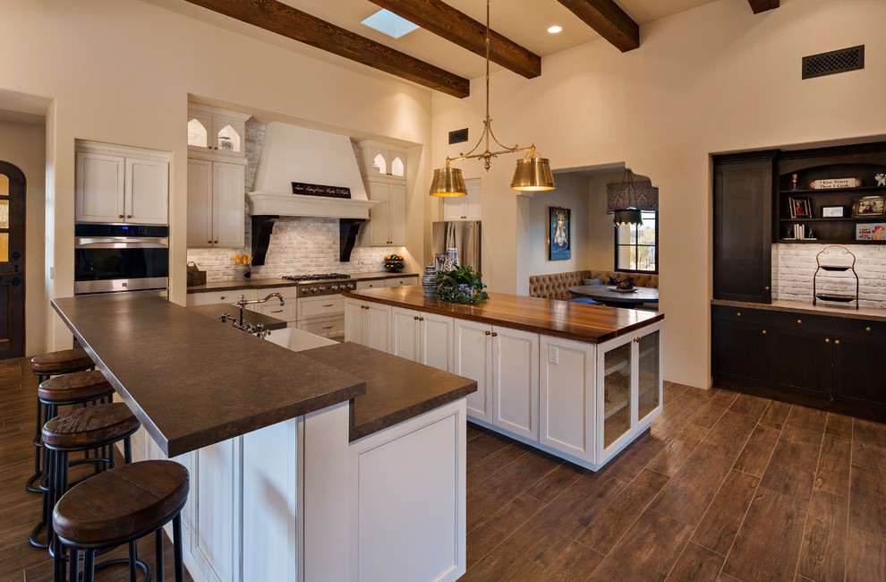 This is an example of a traditional galley open plan kitchen in Phoenix with a farmhouse sink, raised-panel cabinets, white cabinets, quartz benchtops, white splashback, brick splashback, stainless steel appliances, ceramic floors, multiple islands, brown floor and brown benchtop.