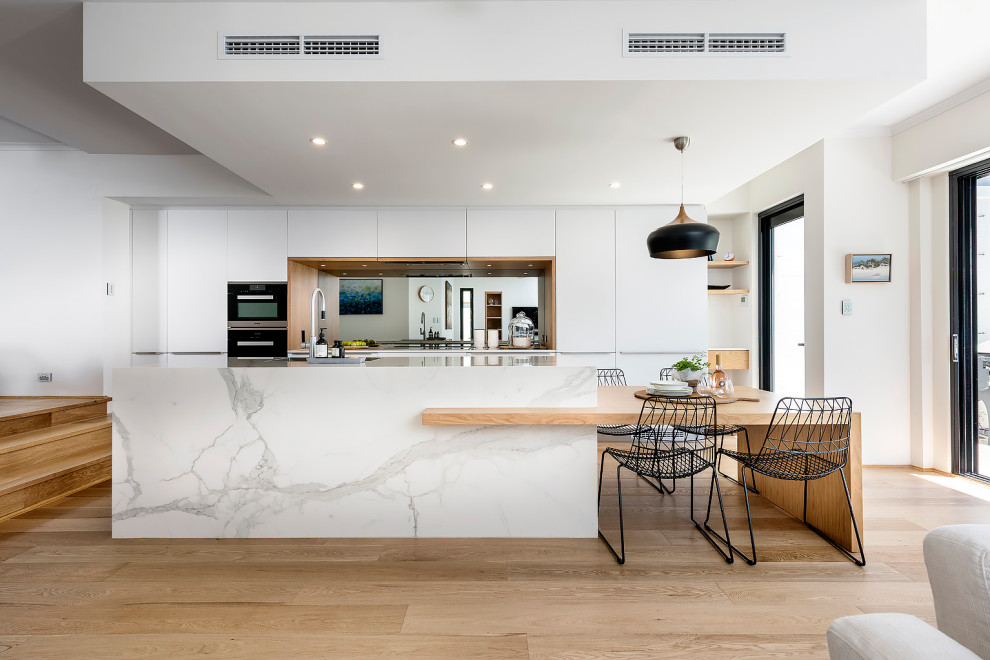 Photo of a large scandinavian galley open plan kitchen in Perth with a single-bowl sink, flat-panel cabinets, white cabinets, quartz benchtops, metallic splashback, mirror splashback, black appliances, light hardwood floors, with island, brown floor and white benchtop.