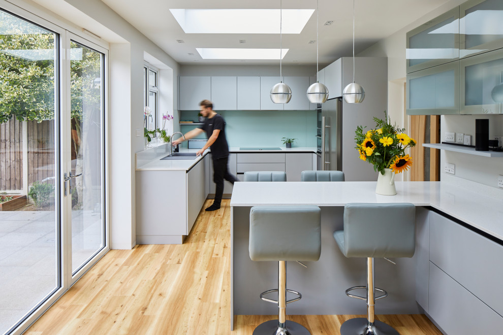 Design ideas for a mid-sized contemporary u-shaped eat-in kitchen in London with flat-panel cabinets, grey cabinets, quartzite benchtops, green splashback, glass sheet splashback, stainless steel appliances, a peninsula and white benchtop.
