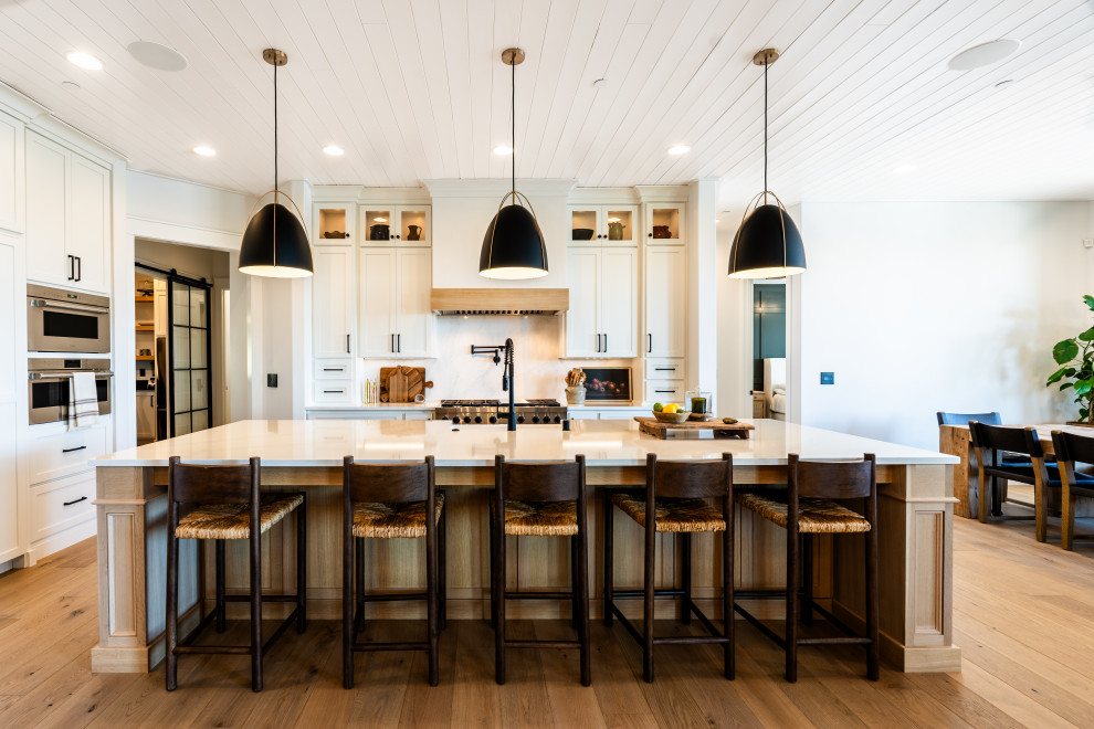This is an example of a transitional l-shaped open plan kitchen in Portland with a farmhouse sink, shaker cabinets, white cabinets, stainless steel appliances, medium hardwood floors, with island, brown floor, white benchtop and timber.
