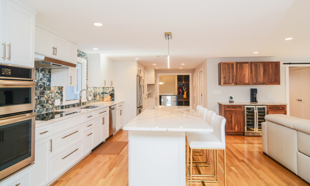 Photo of a large contemporary kitchen/diner in Boston with a single-bowl sink, recessed-panel cabinets, white cabinets, engineered stone countertops, blue splashback, glass tiled splashback, stainless steel appliances, medium hardwood flooring, an island, brown floors and white worktops.