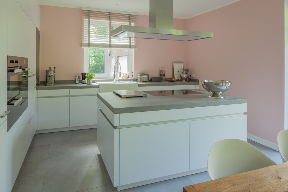 This is an example of a mid-sized contemporary l-shaped open plan kitchen in Hamburg with a farmhouse sink, flat-panel cabinets, white cabinets, wood benchtops, pink splashback, black appliances, porcelain floors, with island and grey floor.
