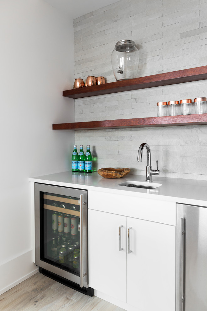 This is an example of a mid-sized beach style single-wall wet bar in New York with an undermount sink, flat-panel cabinets, white cabinets, quartzite benchtops, grey splashback, limestone splashback, light hardwood floors, beige floor and white benchtop.