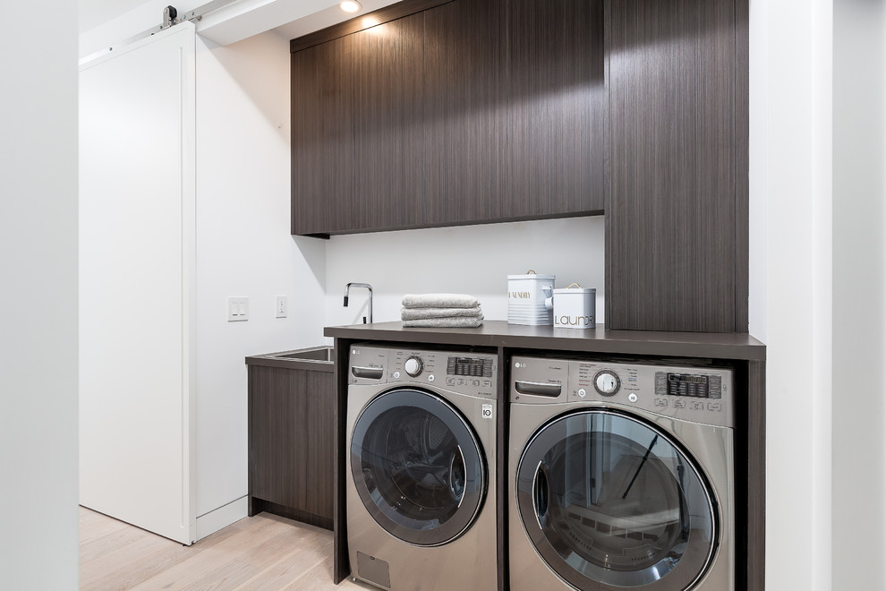 Inspiration for a contemporary single-wall laundry room in Toronto with a single-bowl sink, flat-panel cabinets, dark wood cabinets, white walls, light hardwood floors, a side-by-side washer and dryer and beige floor.
