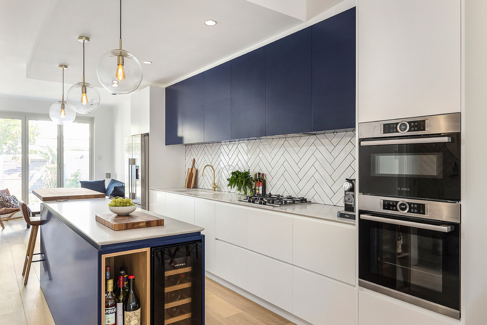 This is an example of a large contemporary galley open plan kitchen in Sydney with an undermount sink, flat-panel cabinets, blue cabinets, white splashback, subway tile splashback, light hardwood floors, with island, white benchtop, quartz benchtops, stainless steel appliances and brown floor.