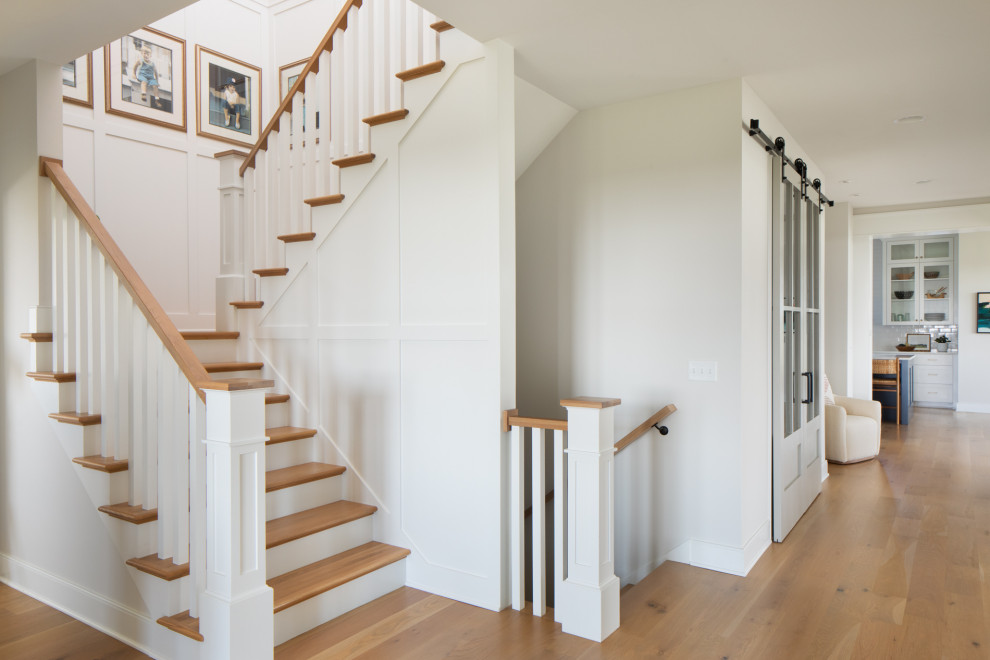 Идея дизайна: большая угловая лестница в стиле неоклассика (современная классика) с деревянными ступенями, крашенными деревянными подступенками, деревянными перилами и панелями на стенах