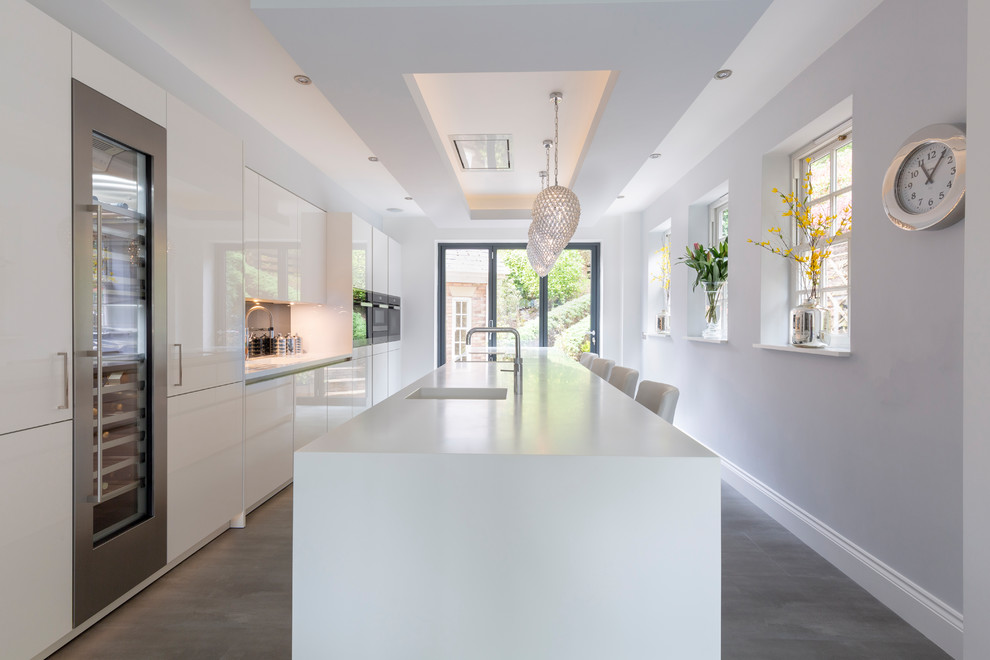 Mid-sized contemporary galley open plan kitchen in Cheshire with flat-panel cabinets, white cabinets, grey splashback, black appliances, porcelain floors, with island, grey floor, white benchtop, an integrated sink, solid surface benchtops and glass sheet splashback.