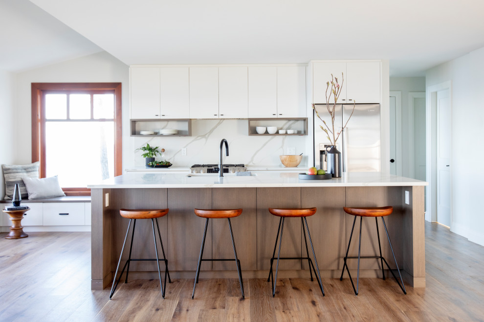 Contemporary galley kitchen in Vancouver with a farmhouse sink, flat-panel cabinets, white cabinets, white splashback, stone slab splashback, stainless steel appliances, medium hardwood floors, with island, brown floor and white benchtop.