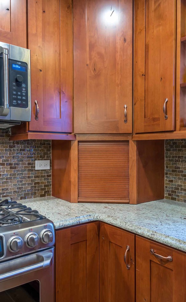 This is an example of a mid-sized country l-shaped eat-in kitchen in Chicago with an undermount sink, shaker cabinets, medium wood cabinets, granite benchtops, brown splashback, glass tile splashback, stainless steel appliances, ceramic floors, with island, brown floor, grey benchtop and wallpaper.