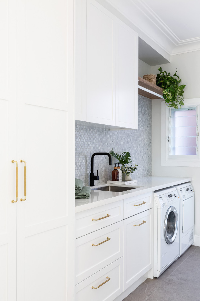 Design ideas for a mid-sized beach style dedicated laundry room in Sydney with a single-bowl sink, shaker cabinets, white cabinets, quartzite benchtops, marble splashback, white walls, porcelain floors, a side-by-side washer and dryer, white benchtop, grey splashback and grey floor.