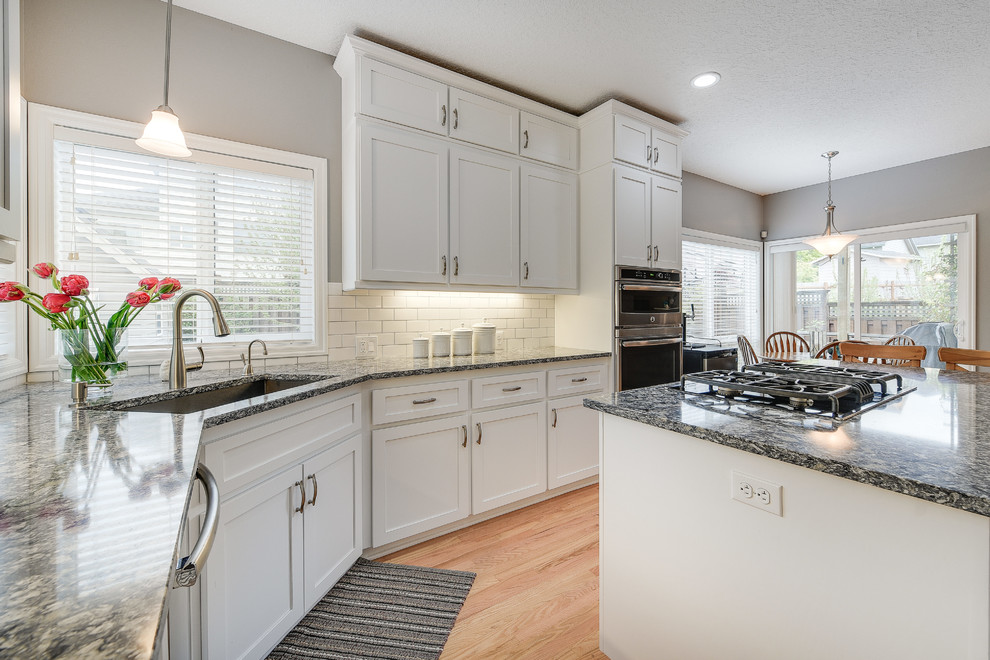 Example of a mid-sized transitional l-shaped light wood floor and brown floor enclosed kitchen design in Portland with a drop-in sink, shaker cabinets, white cabinets, quartz countertops, white backsplash, subway tile backsplash, an island and stainless steel appliances