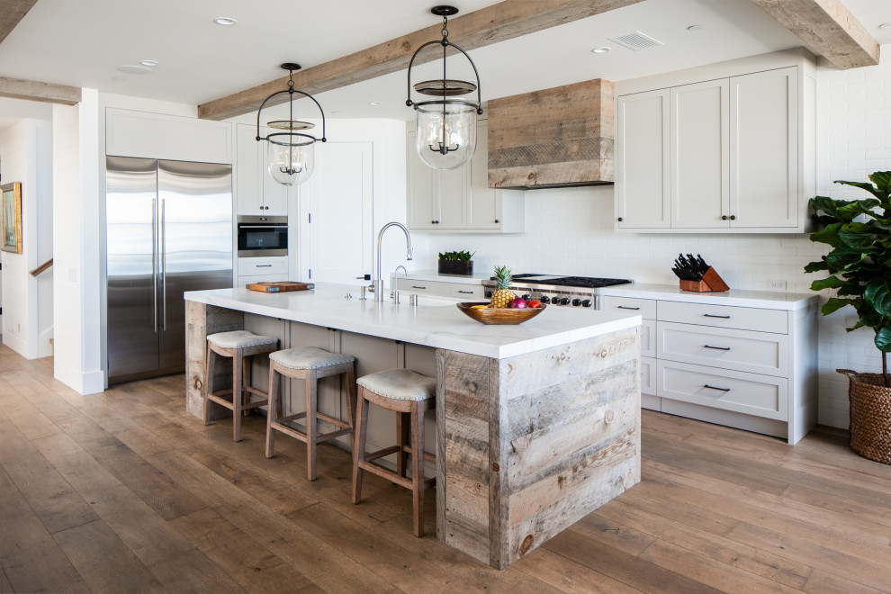 Photo of a mid-sized beach style open plan kitchen in Orange County with an undermount sink, shaker cabinets, white cabinets, marble benchtops, white splashback, ceramic splashback, stainless steel appliances, medium hardwood floors, with island and white benchtop.