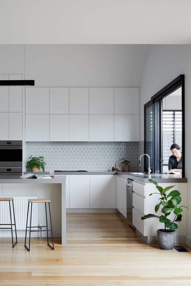 Design ideas for a large contemporary l-shaped open plan kitchen in Melbourne with a double-bowl sink, flat-panel cabinets, white cabinets, quartz benchtops, white splashback, ceramic splashback, stainless steel appliances, light hardwood floors, with island, brown floor and grey benchtop.